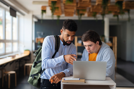 A male tutor, wearing a backpack, gestures toward the screen while explaining something to a man with down syndrome wearing a hoodie, listens closely. A notebook and markers rest on the table in front of them.
